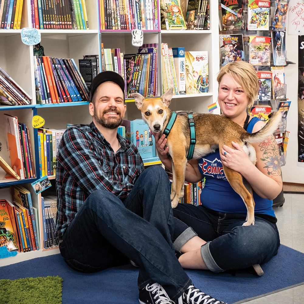 Sarah & Patrick Titus with their rescue dog, Heyzel. They are smiling and sitting on a colored mat in front of bookcases of graphic novels for younger readers.